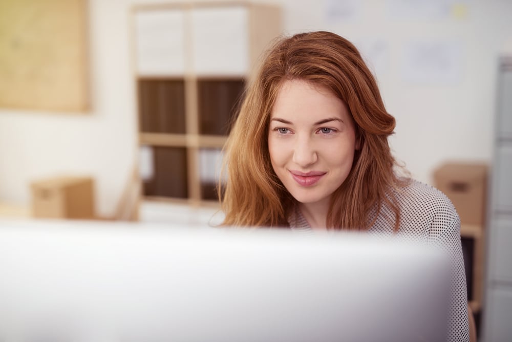 Attractive young woman working on a desktop computer smiling as she leans forwards reading text on the screen, view over the monitor Attractive young woman working on a desktop computer smiling as she leans forwards reading text on the screen, view over the monitor