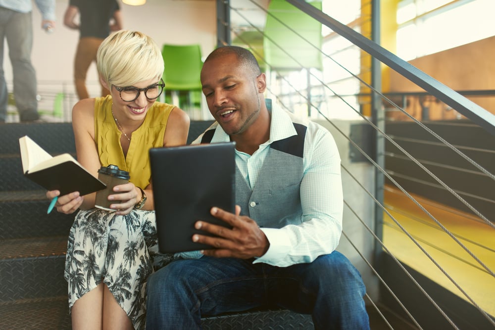 Two creative millenial small business owners working on social media strategy using a digital tablet while sitting in staircase Two creative millenial small business owners working on social media strategy using a digital tablet while sitting in staircase